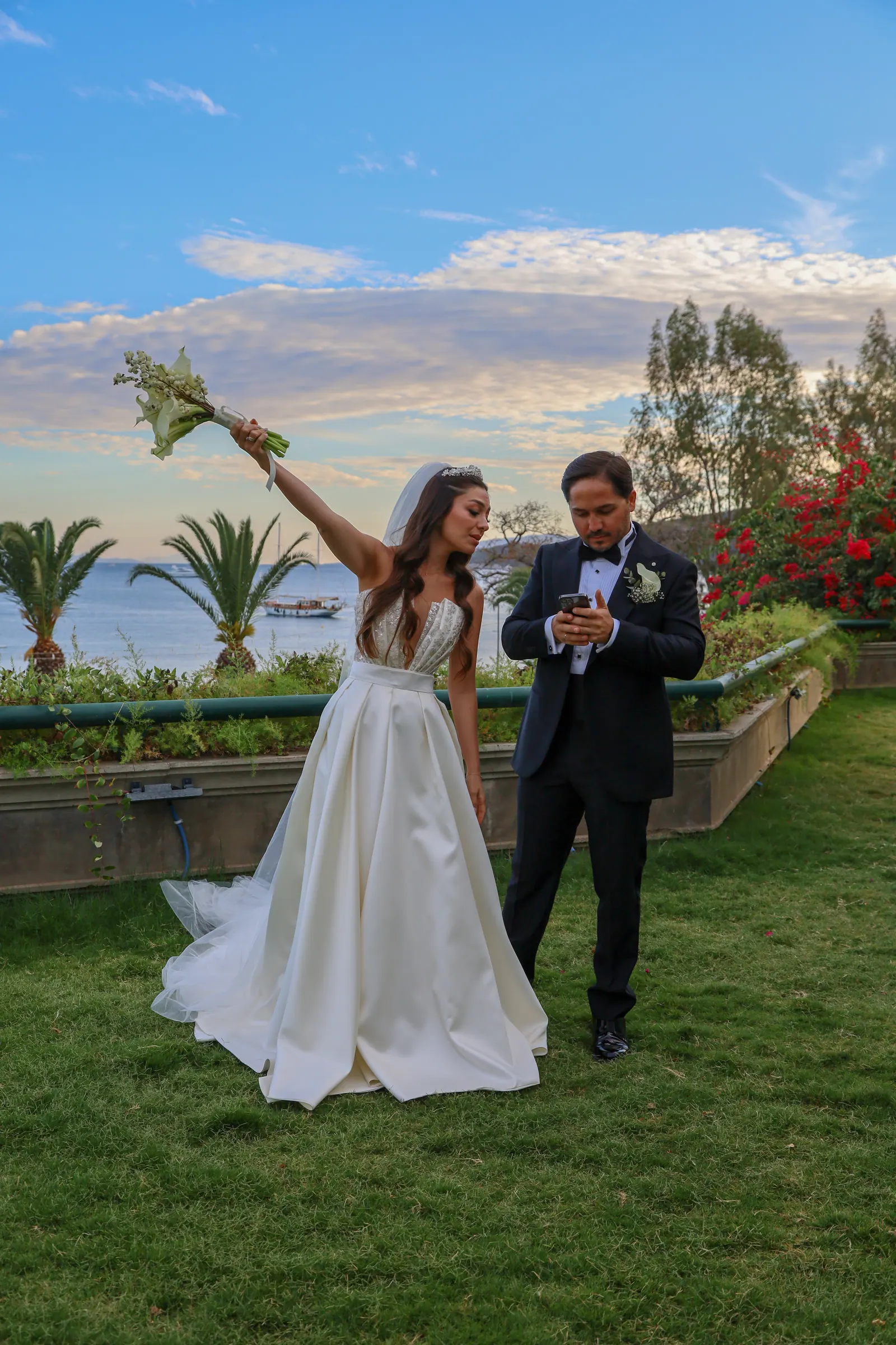 Deniz manzaralı bahçede buketini havaya kaldıran gelin ve damat, koyda gulet