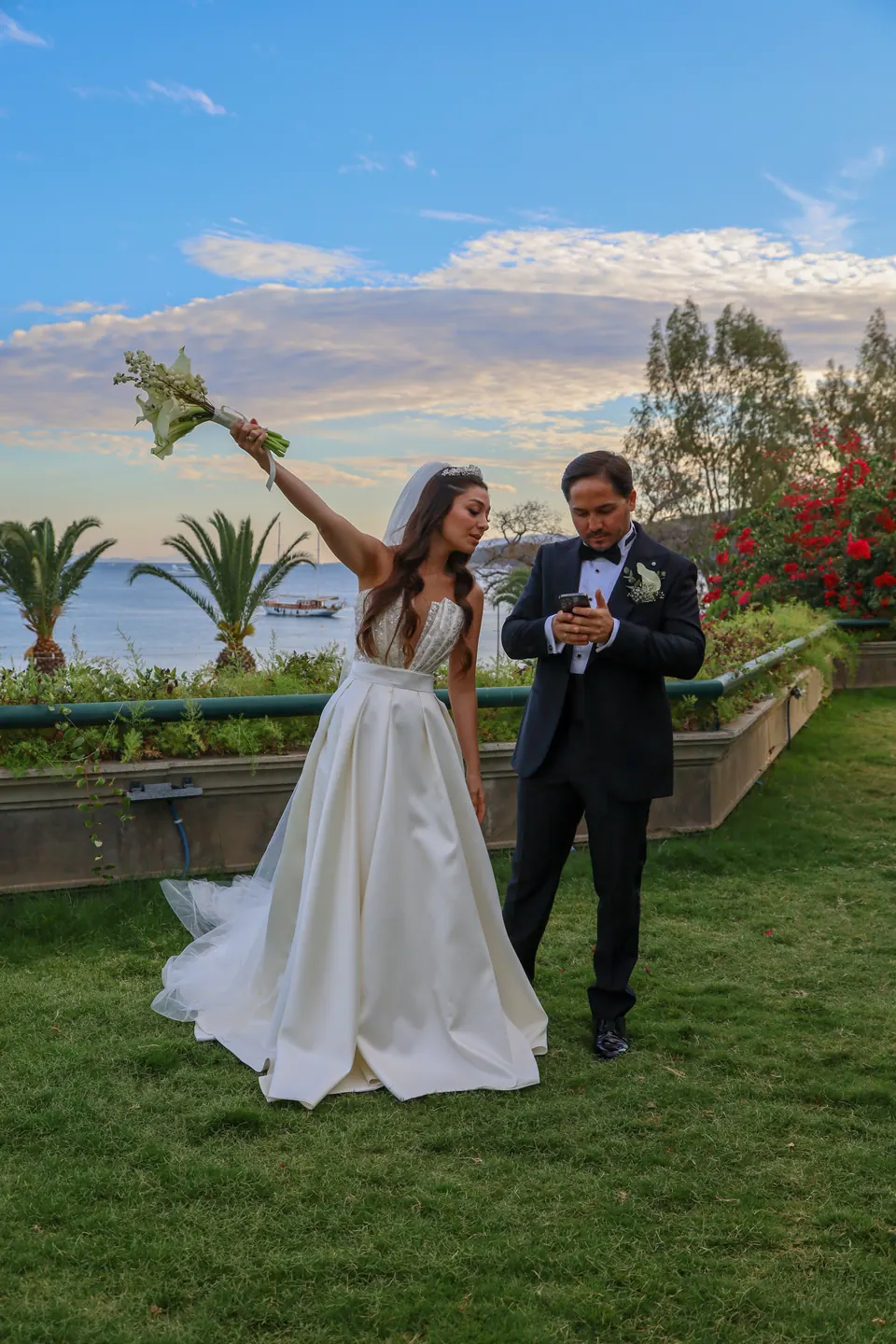 Deniz manzaralı bahçede buketini havaya kaldıran gelin ve damat, koyda gulet