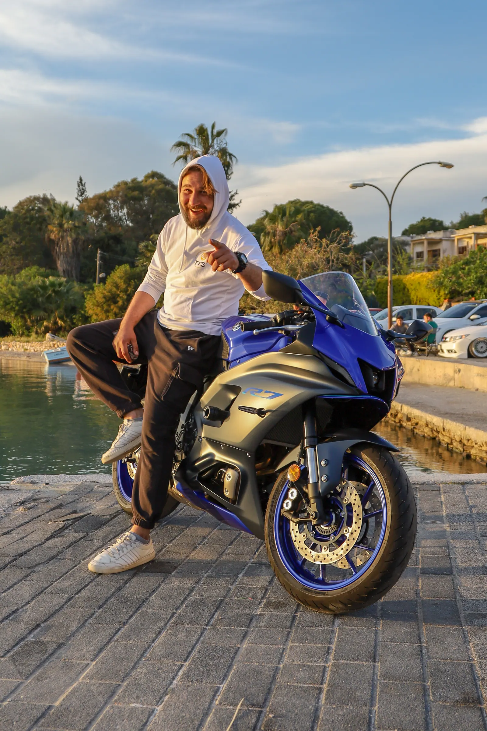 Gün batımında liman kenarında spor motosiklete yaslanan gülümseyen adam