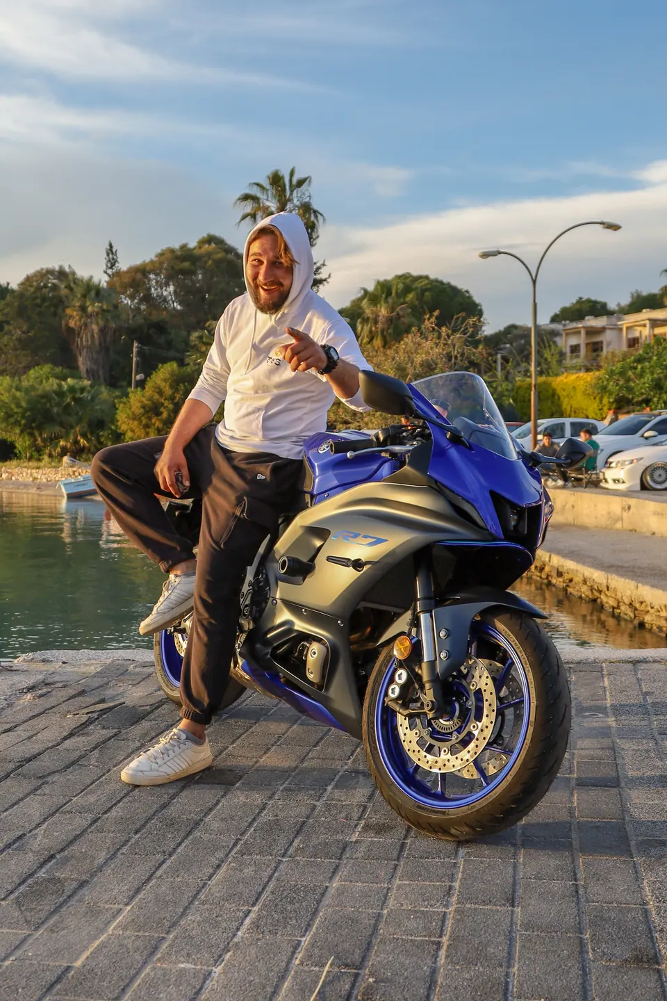Gün batımında liman kenarında spor motosiklete yaslanan gülümseyen adam
